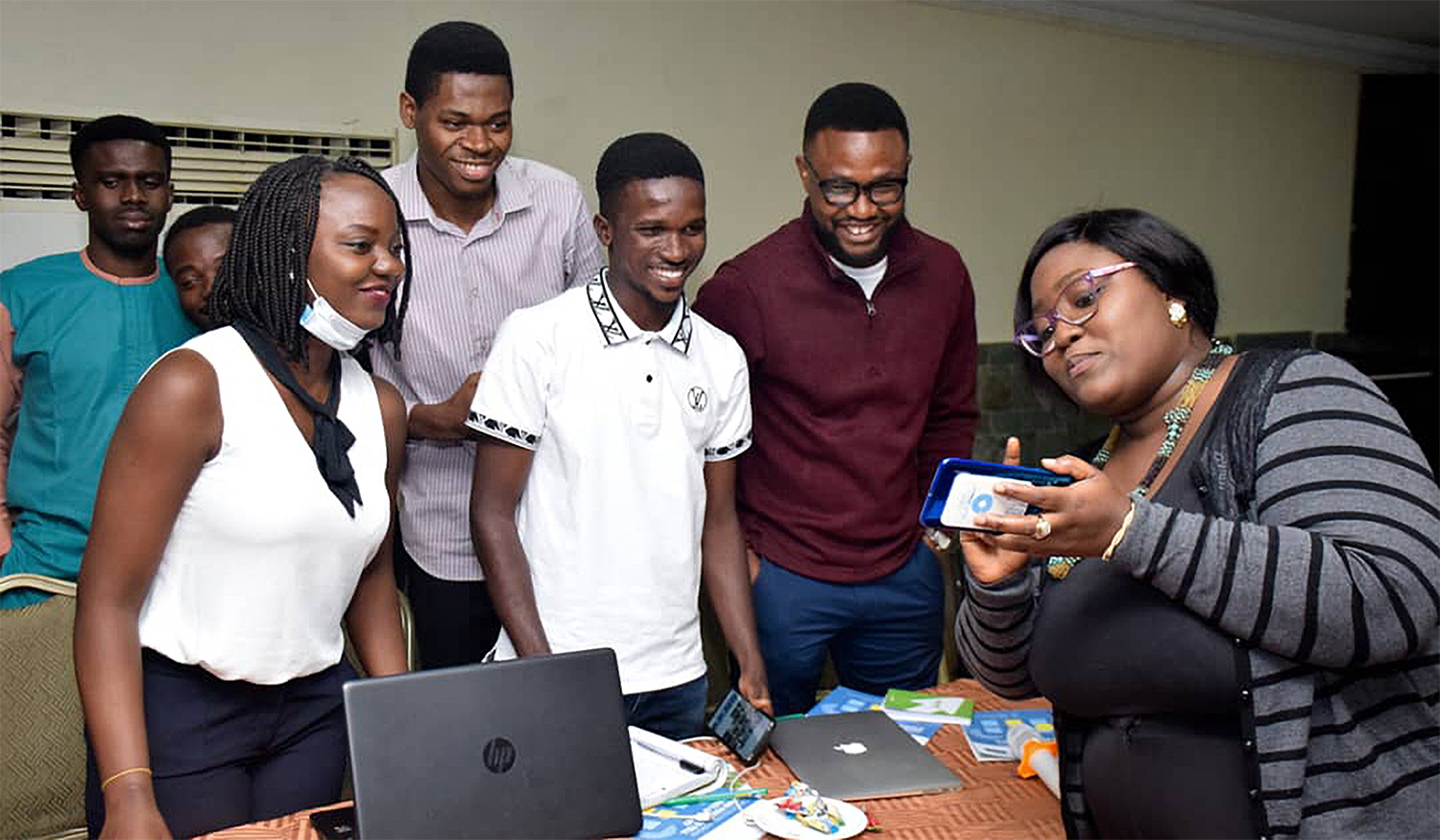 A group of people smiling and looking at a phone, with laptops and papers on a table in front of them.